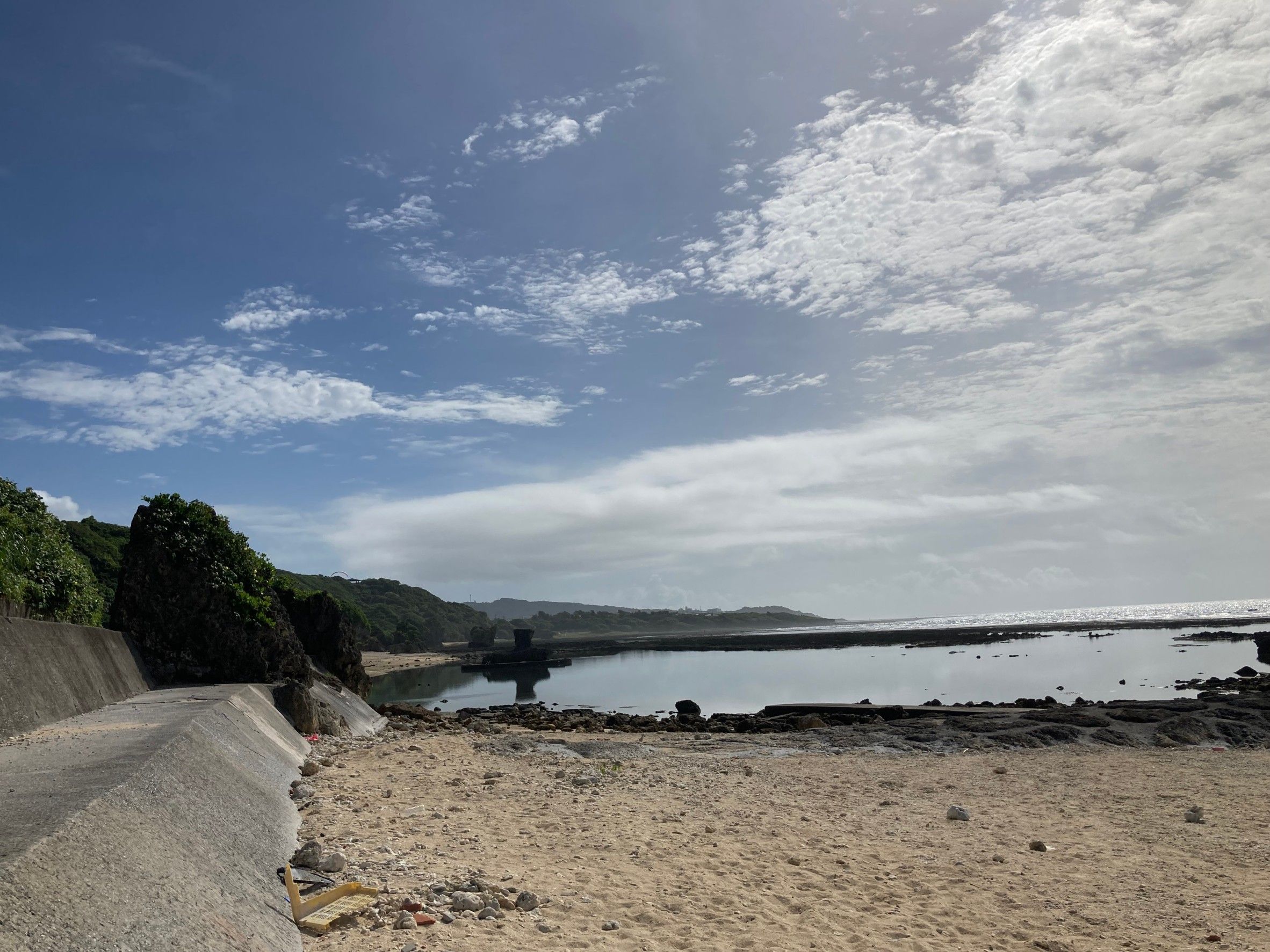 A coastal landscape with a sandy beach, rocky shoreline, and a calm sea under a partly cloudy sky.
