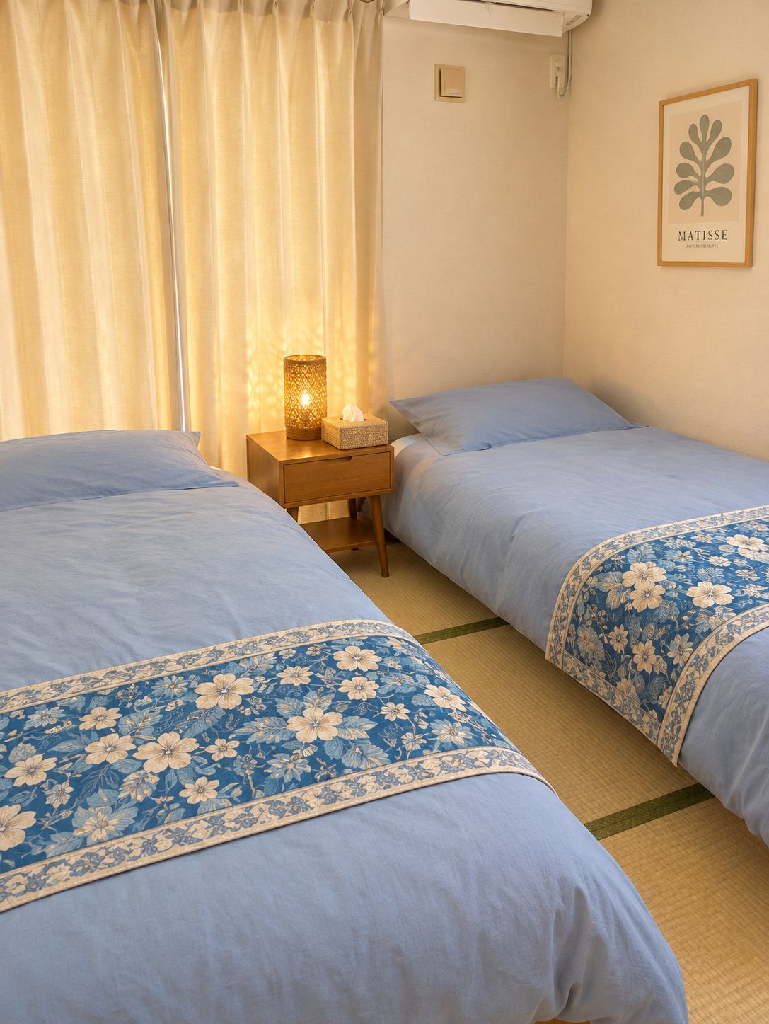 A hotel room with two beds, a bedside table with a lamp, a framed artwork on the wall, and a window with curtains.