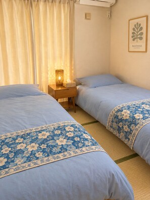 A hotel room with two beds, a bedside table with a lamp, a framed artwork on the wall, and a window with curtains.