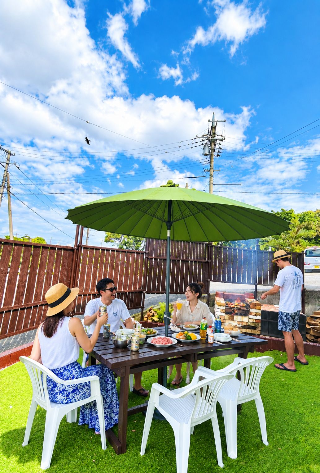 Un grupo de personas cenando al aire libre bajo un paraguas verde, con una barbacoa en el fondo.