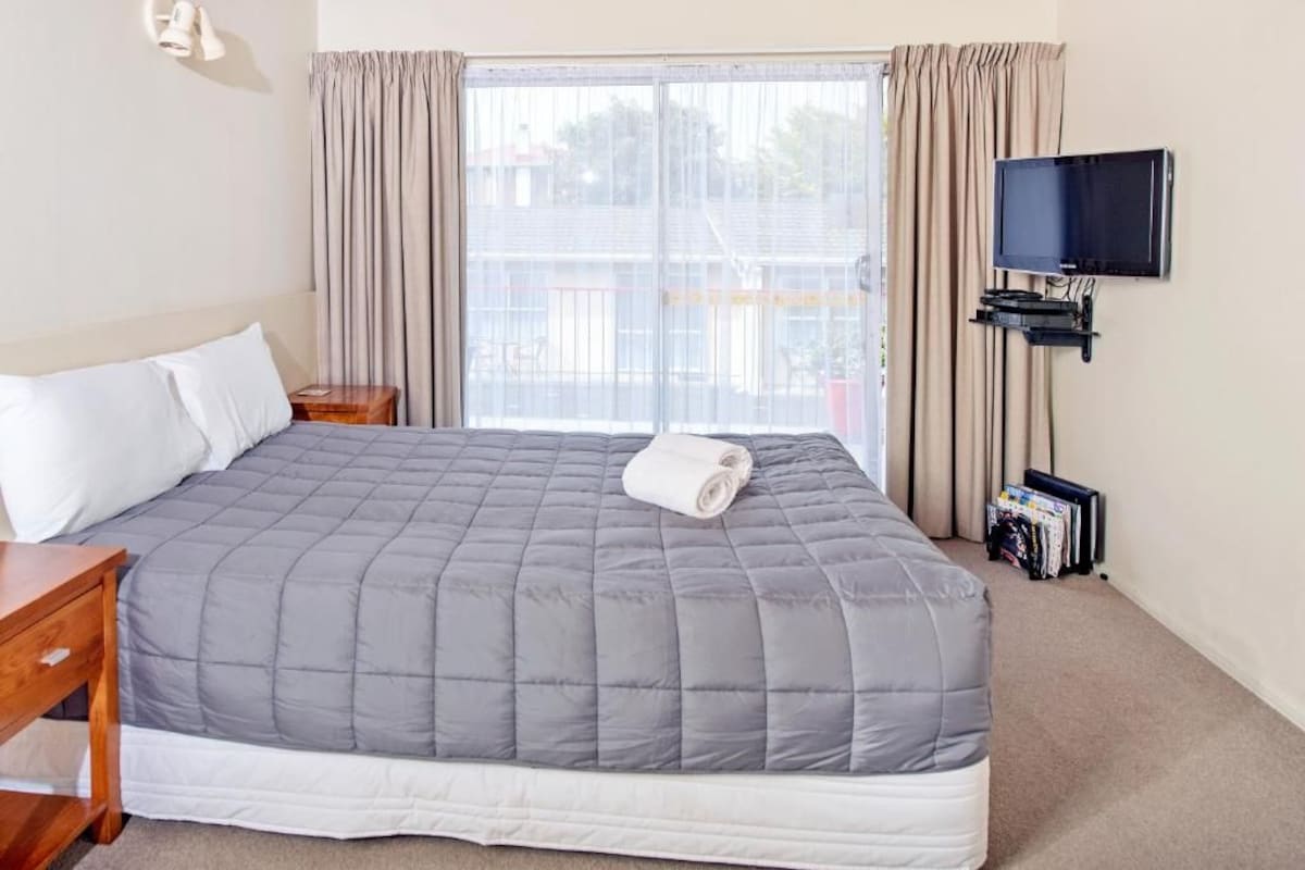 Habitación de hotel con una cama grande, un televisor montado en la pared y una ventana con cortinas.
