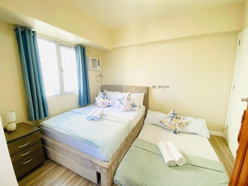 A hotel room with two beds, a bedside table, a window with curtains, and a wall-mounted air conditioner.