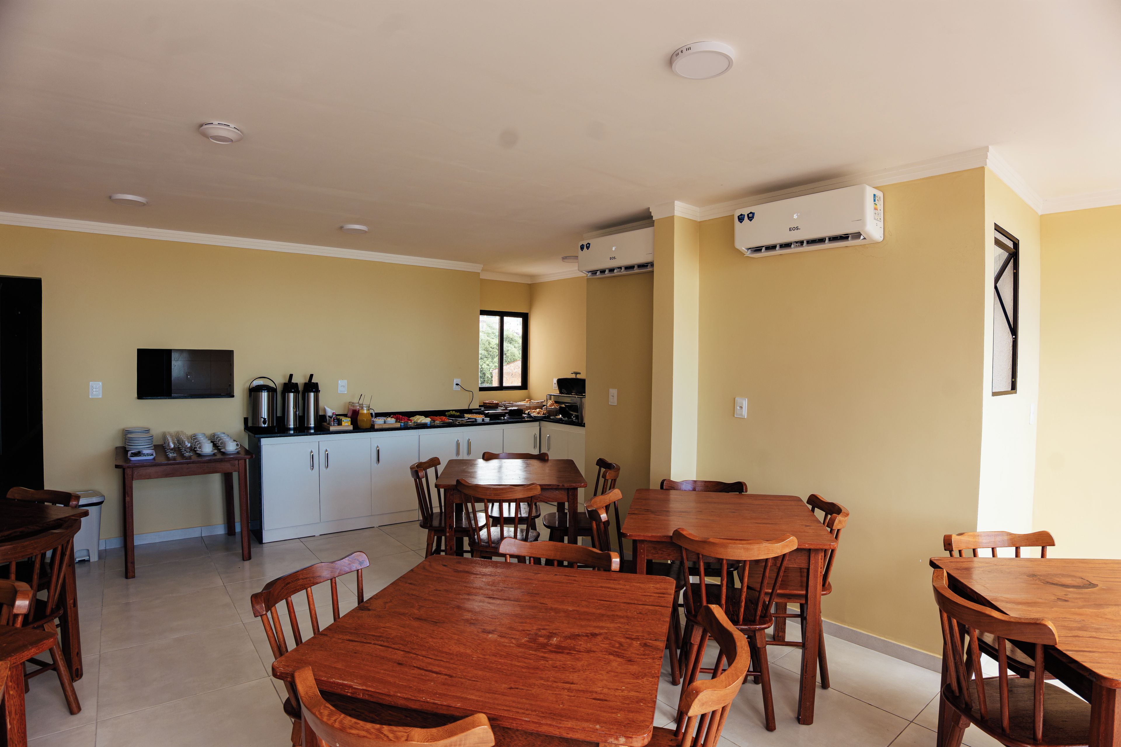 A dining area with wooden tables and chairs, a kitchen with a window, and an air conditioning unit on the wall.