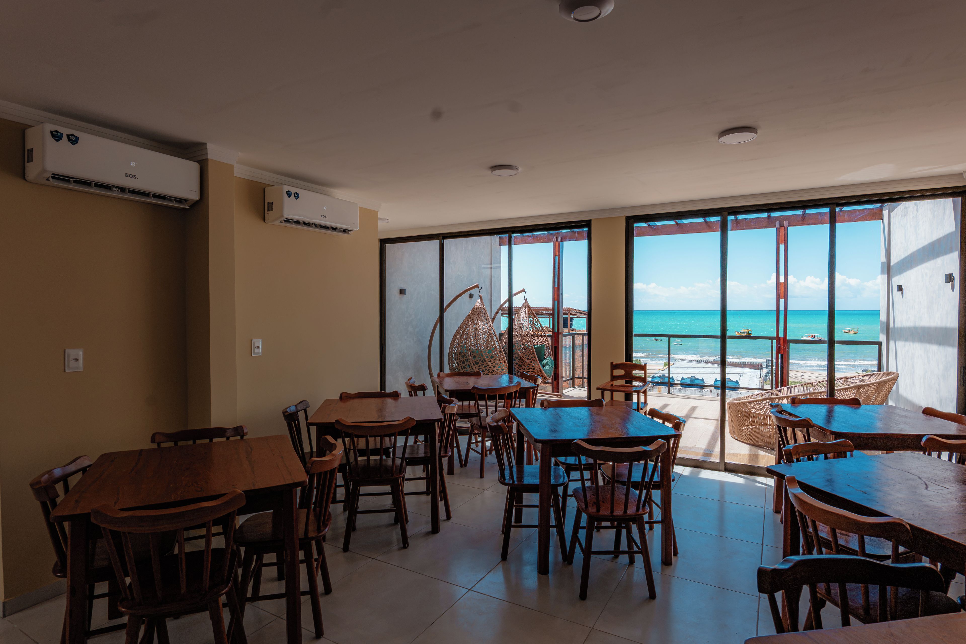 A restaurant with wooden tables and chairs, large windows with a view of the beach and ocean, and air conditioning units on the wall.