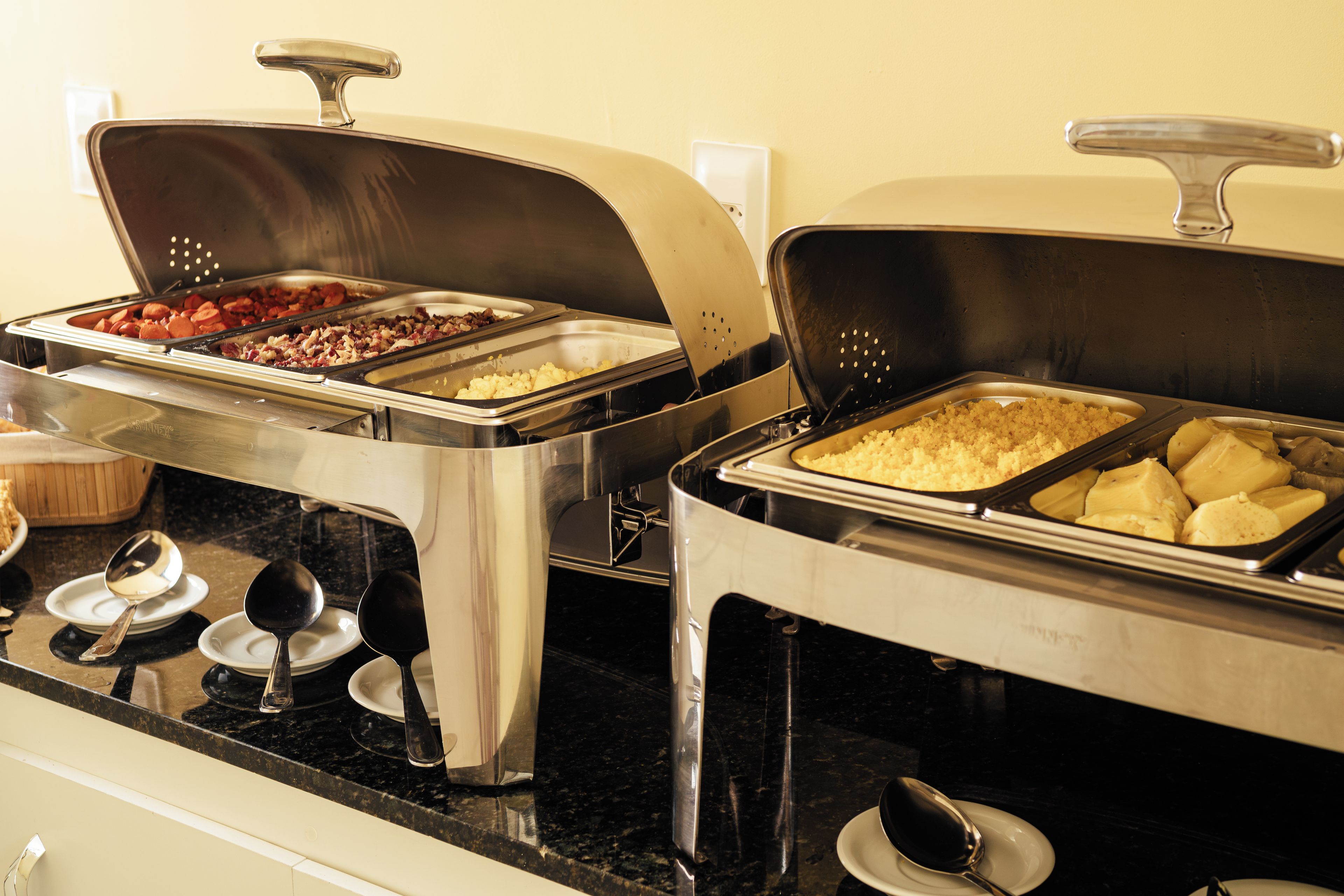 A buffet setup with various food items in metal serving trays, a spoon on a plate, and a black countertop.