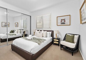A modern bedroom with a large bed, bedside tables, a chair, and a mirror. - Salty Air Coastal Cottage, Pet Friendly (Lake Illawarra)