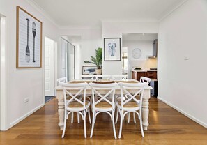 A dining area with a white table and chairs, a clock on the wall, and a plant in the background. - Salty Air Coastal Cottage, Pet Friendly (Lake Illawarra)