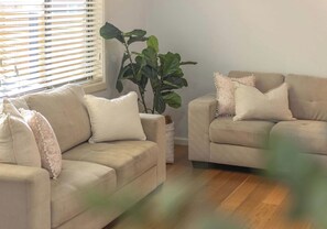 A living room with two beige sofas, one with a potted plant, and a window with blinds. - Salty Air Coastal Cottage, Pet Friendly (Lake Illawarra)