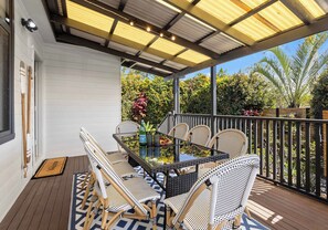 A covered outdoor patio with a glass-top table and wicker chairs, surrounded by a wooden deck and a metal railing. - Salty Air Coastal Cottage, Pet Friendly (Lake Illawarra)