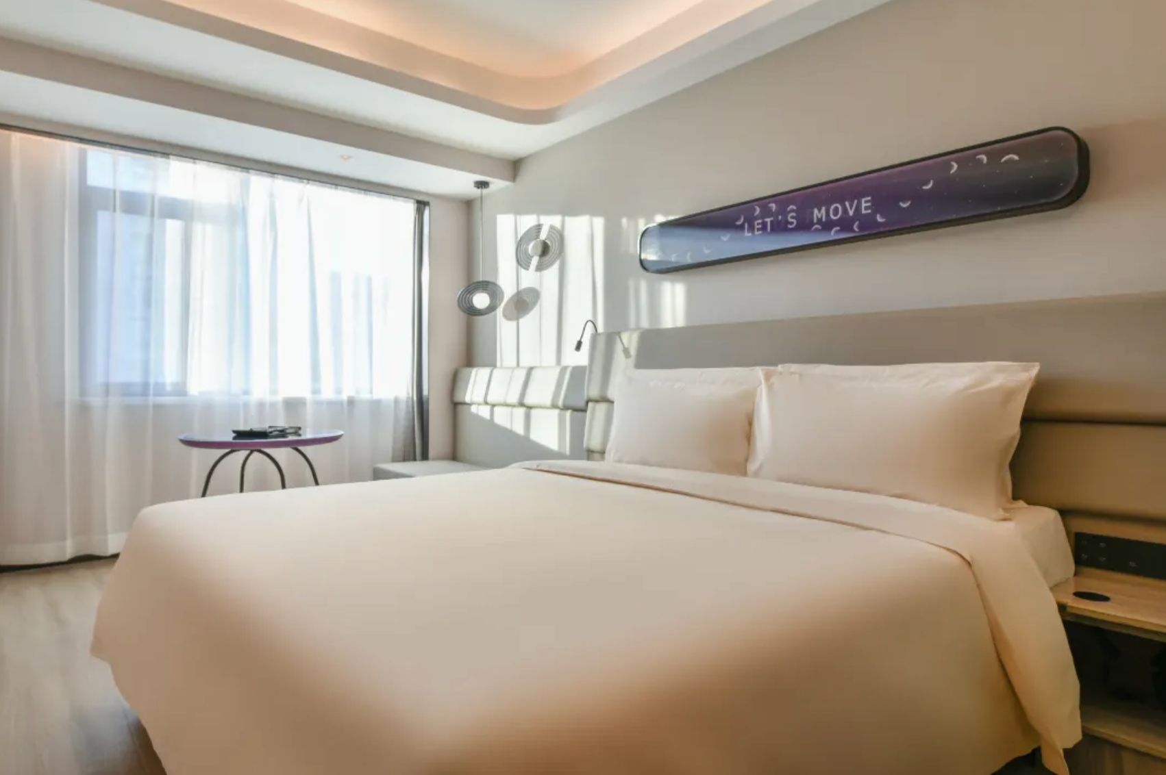 A hotel room with a large bed, a bedside table, a small table, and a wall-mounted light fixture.