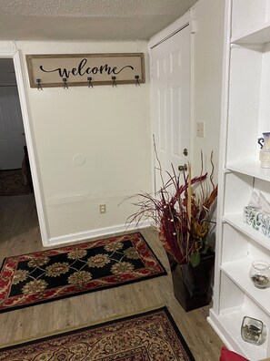 A hallway with a welcome sign, a doormat, a vase with dried plants, and a shelf with decorative items. - Cozy and Comfy "Away from Home" (Pine Bluff)