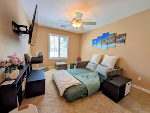 Ein Schlafzimmer mit Bett, Nachttischen, Schreibtisch, Deckenventilator, Fenster mit Jalousien und Wandbildern.