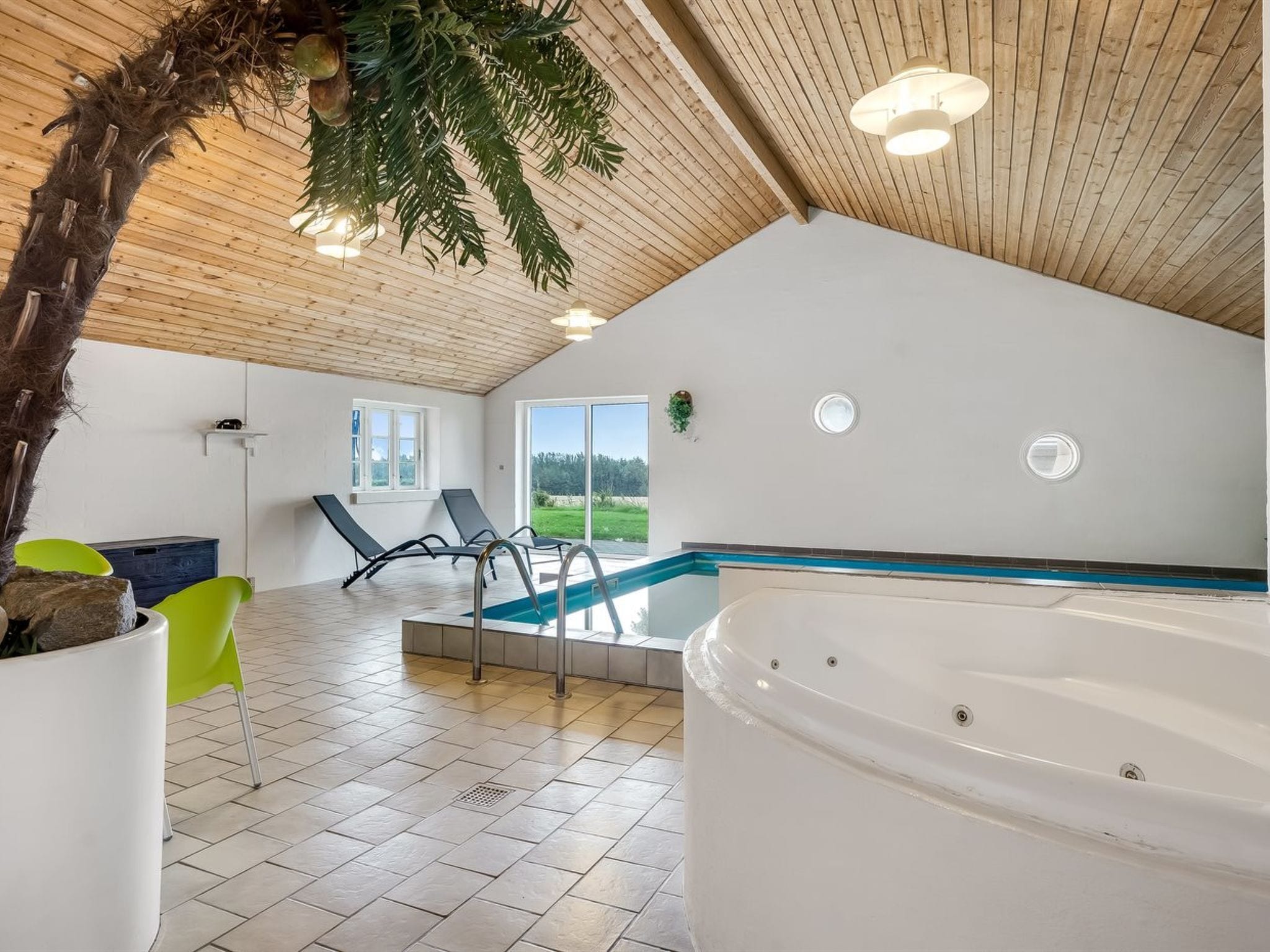 A modern indoor swimming pool with a wooden ceiling, two lounge chairs, and a jacuzzi.