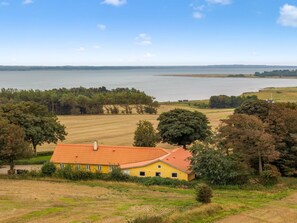 Een geel gebouw met een rood dak, omringd door velden en bomen, met uitzicht op een groot wateroppervlak.
