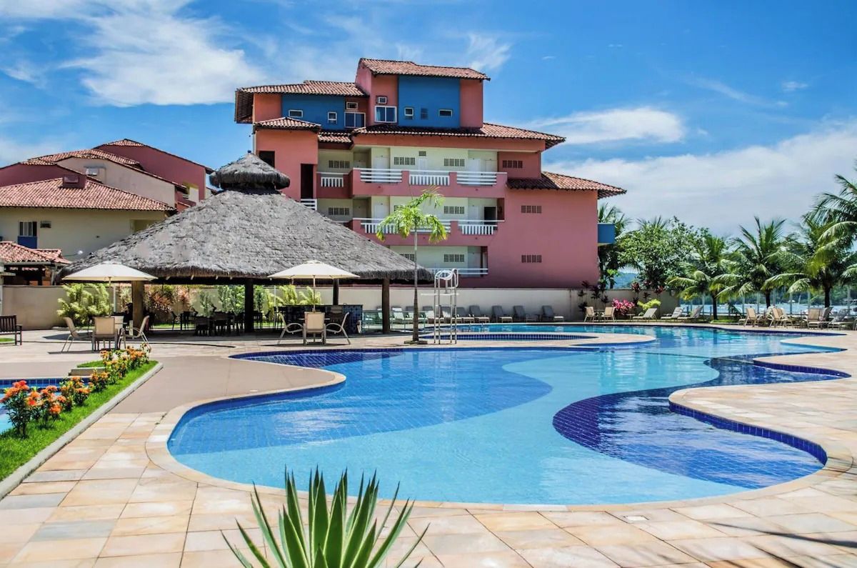 A swimming pool with lounge chairs and umbrellas, surrounded by buildings with balconies and a clear sky.