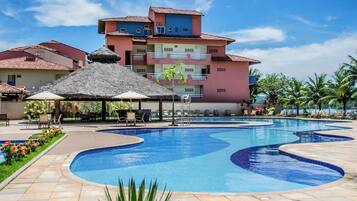 A swimming pool with lounge chairs and umbrellas, surrounded by buildings with balconies and a clear sky.