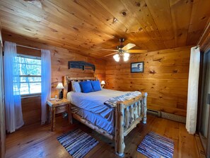 A wooden cabin bedroom with a bed, nightstands, a ceiling fan, and a window with curtains.