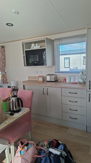 A compact kitchen with a microwave, toaster, and kettle on the counter, a small table with a chair, and a window with blinds.