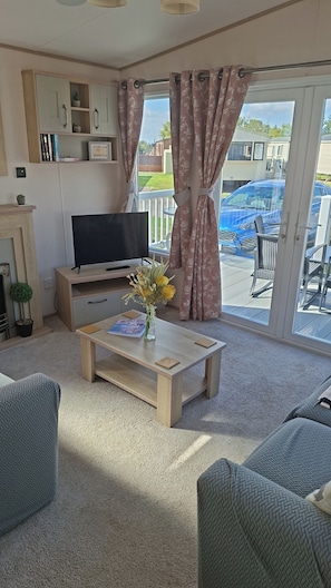 A living room with a TV, a coffee table with a vase, and a view of a blue car outside. - 30C Kingfisher Hottub Lodge (Tattershall)