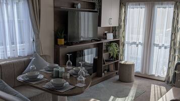 A modern living room with a flat-screen TV, a sofa, a dining table set for four, and a large window with sheer curtains.