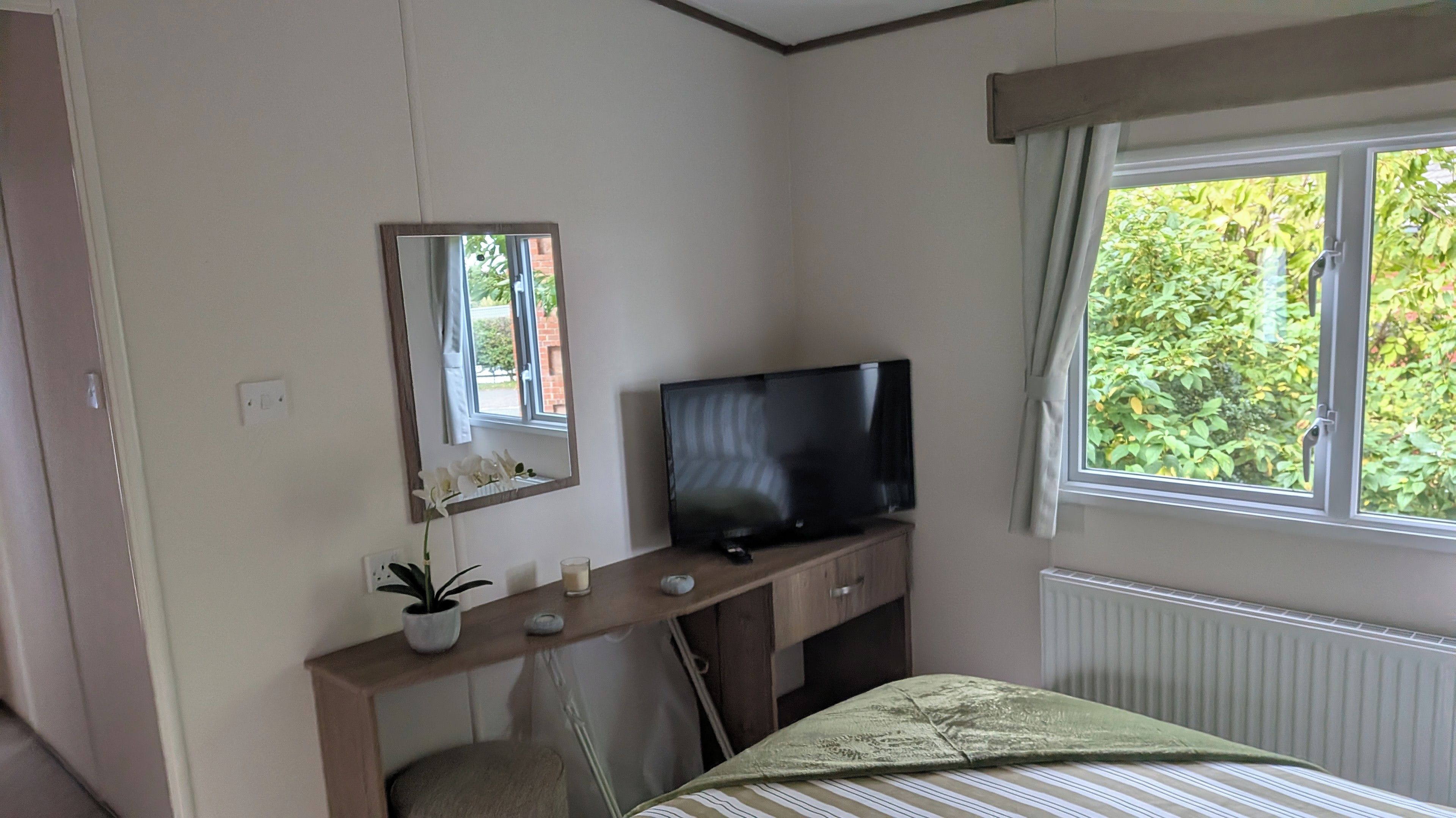 A hotel room with a bed, a TV on a wooden dresser, a mirror, and a window with a view of greenery.