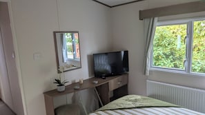 A hotel room with a bed, a TV on a wooden dresser, a mirror, and a window with a view of greenery. - Caravan sleeps 6 with access to indoor pool (Dunbar)
