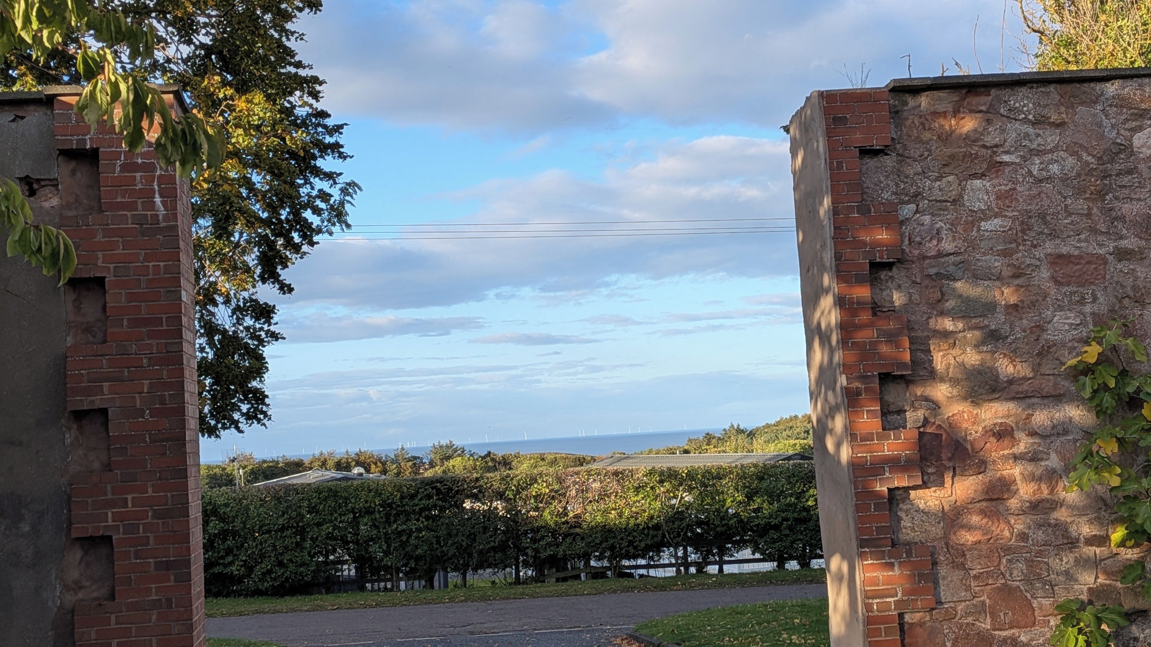 A brick wall with a gate leading to a landscaped area with a hedge and a view of wind turbines in the distance.