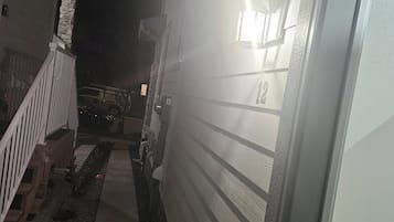 Un sendero estrecho que conduce a un edificio con una puerta blanca, un muro de piedra y un vehículo estacionado afuera.