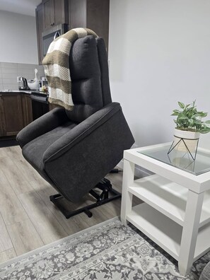 Una sala moderna con un sillón reclinable gris, una mesita auxiliar blanca con una planta y una alfombra estampada.