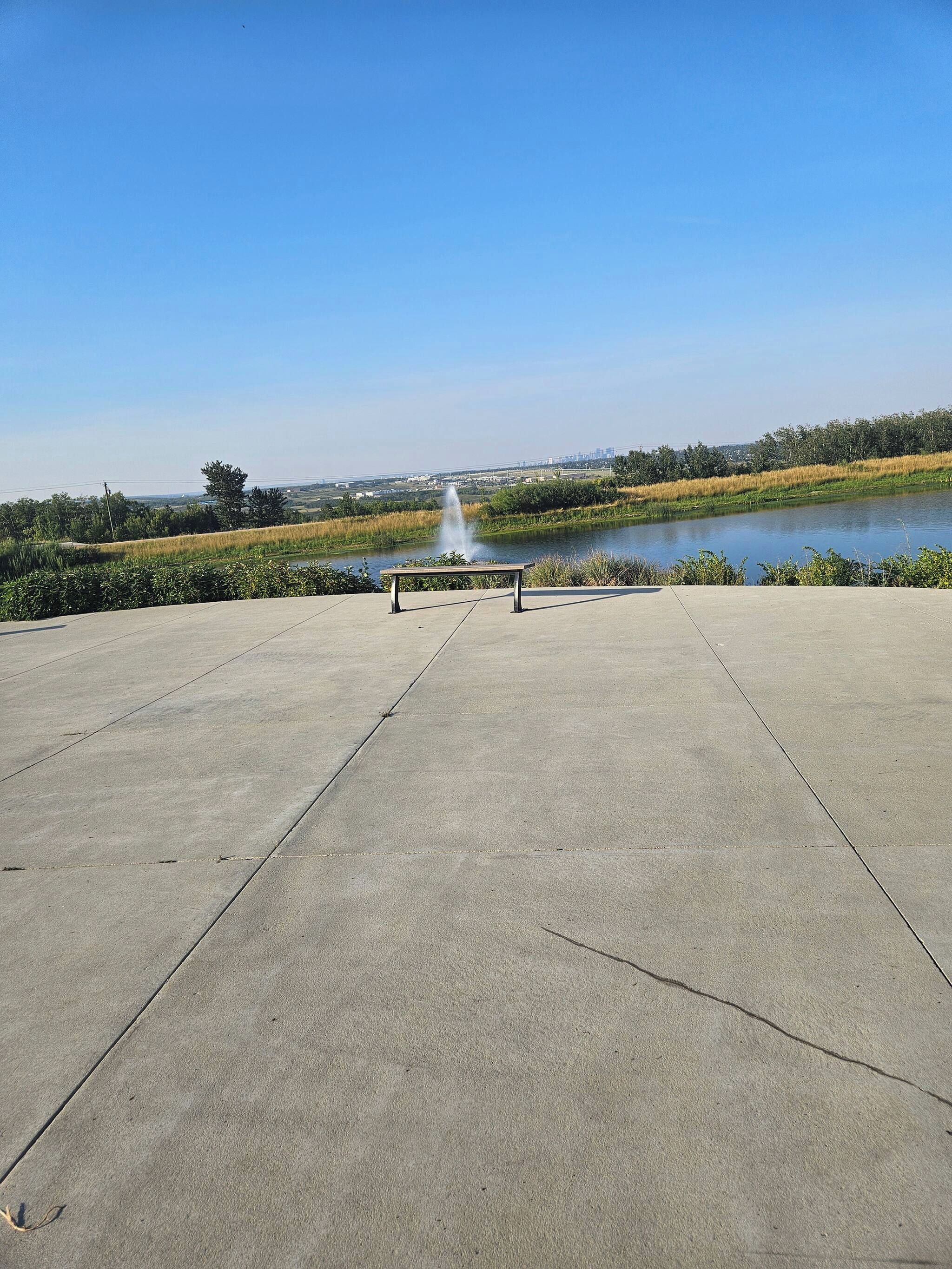Eine Beton-Aussichtsplattform mit einer Bank und einem Springbrunnen in der Mitte, mit Blick auf ein Gewässer und eine entfernte Landschaft.