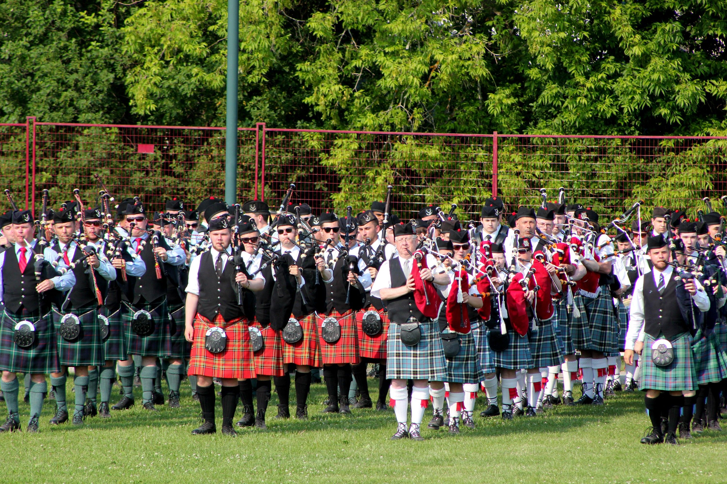 Un groupe de personnes, vêtues de costumes traditionnels écossais, notamment des kilts et des cornemuses, se tient en rangée à l’extérieur.