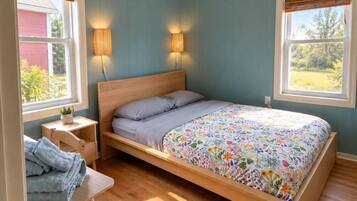 A bedroom with a single bed, bedside lamps, a small table with a chair, and a window with a view of greenery.
