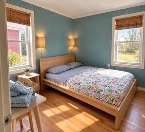 A bedroom with a single bed, bedside lamps, a small table with a chair, and a window with a view of greenery.
