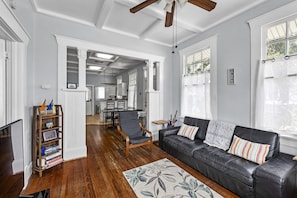 A spacious living room with a black leather sofa, a wooden bookshelf, and a dining area in the background. - Galvie House | Walk to Beach + Restaurants (Galveston)