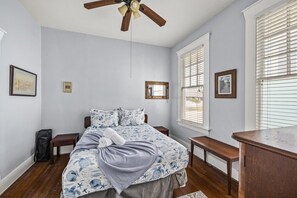 Une chambre avec un lit, un ventilateur de plafond, une fenêtre avec des stores et un bureau.