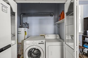 Une petite buanderie équipée de deux machines à laver, d’un chauffe-eau et d’une étagère sur laquelle se trouve une bouteille de lessive.