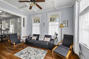 Ein Wohnzimmer mit einer schwarzen Ledersofa, zwei Sesseln, einem Holztisch, einem gemusterten Teppich und einem Deckenventilator.