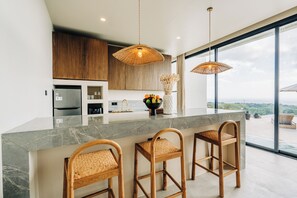 A modern kitchen with a marble countertop, wooden cabinets, and a large window offering a view. - Villa Veluna Uluwatu (Uluwatu)