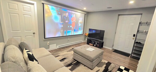 Una sala de estar con un televisor de pantalla plana grande, un sofá, un reposapiés y piso alfombrado.