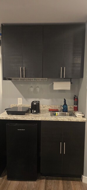 Une cuisine de taille réduite, avec des meubles de couleur sombre, un grille-pain, un distributeur d’eau et un extincteur.