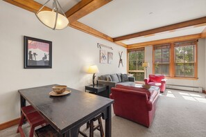 Una sala de estar con una mesa de madera, un sofá rojo y un sofá gris. Hay un cartel enmarcado en la pared y una ventana con vistas a árboles.