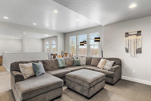 A modern living room with a sectional sofa, ottoman, and a dining area with a table and chairs. - 4BR, 4BA villa in Sienna Hills with pool, hot tub & resort amenities. (Washington)
