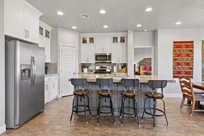 A modern kitchen with white cabinetry, a stainless steel refrigerator, a microwave, and a granite countertop. There are four bar stools with wooden seats and black metal frames. - 4BR, 4BA villa in Sienna Hills with pool, hot tub & resort amenities. (Washington)