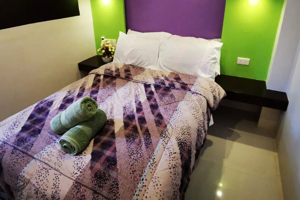 Una cama bien hecha con un edredón estampado y dos toallas enrolladas, una mesita de noche con una lámpara, y una pared con un panel morado e iluminación verde.