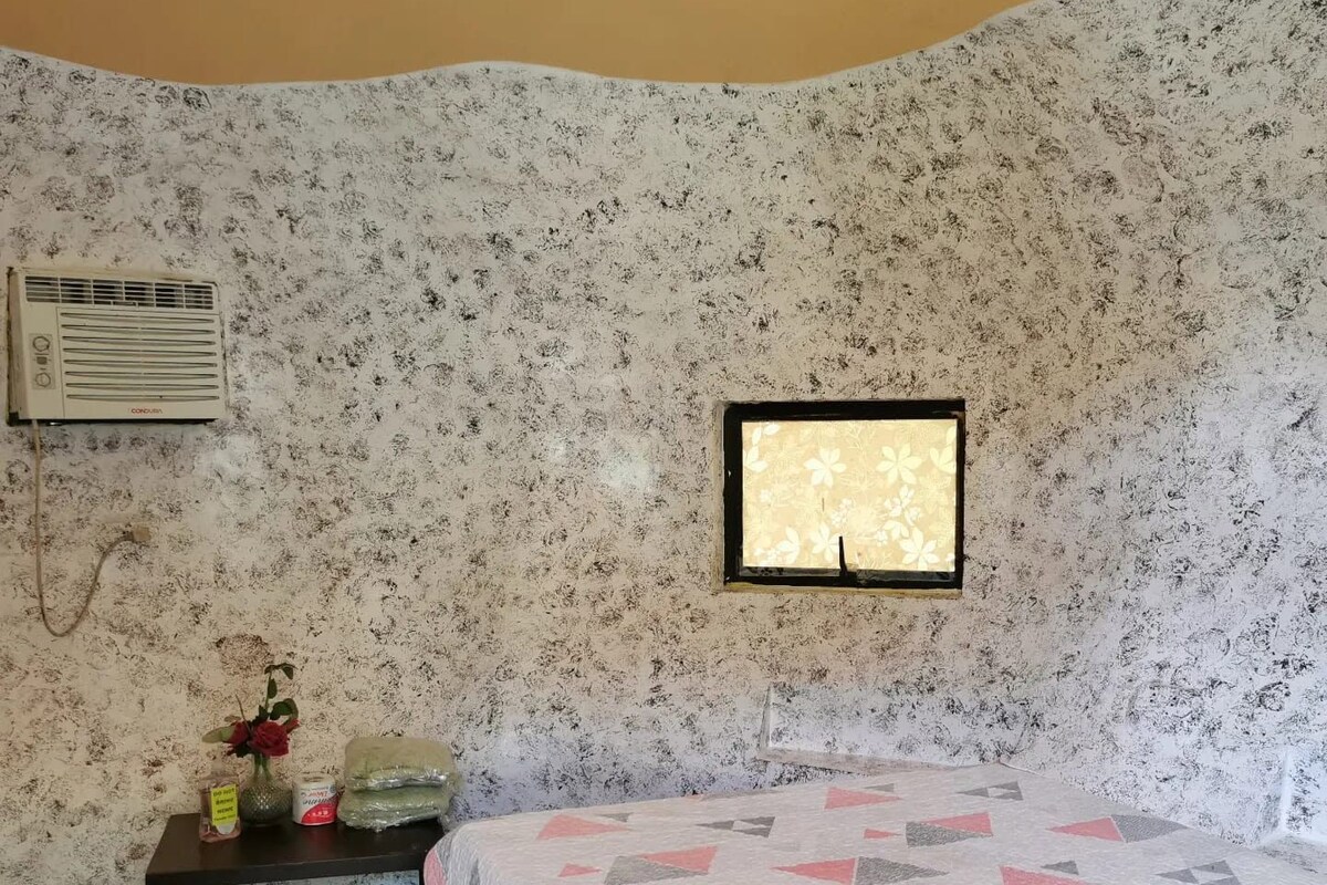 Una habitación con una cama, una mesita con un jarrón de flores, un aire acondicionado instalado en la pared y una ventana con estampado floral.