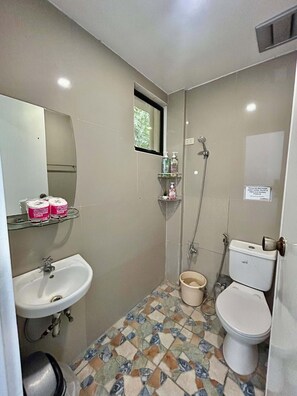 Un baño pequeño con lavamanos, inodoro y ducha. Hay una ventana sobre el espejo y un cartel en la pared.