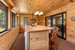 A wooden cabin interior with a central island, large windows, and a dining area.