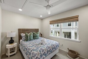 A bedroom with a bed, a nightstand, a lamp, a window with a view of buildings, and a ceiling fan. - Casa Flamingo- Canal Front | Great for Families (Galveston)