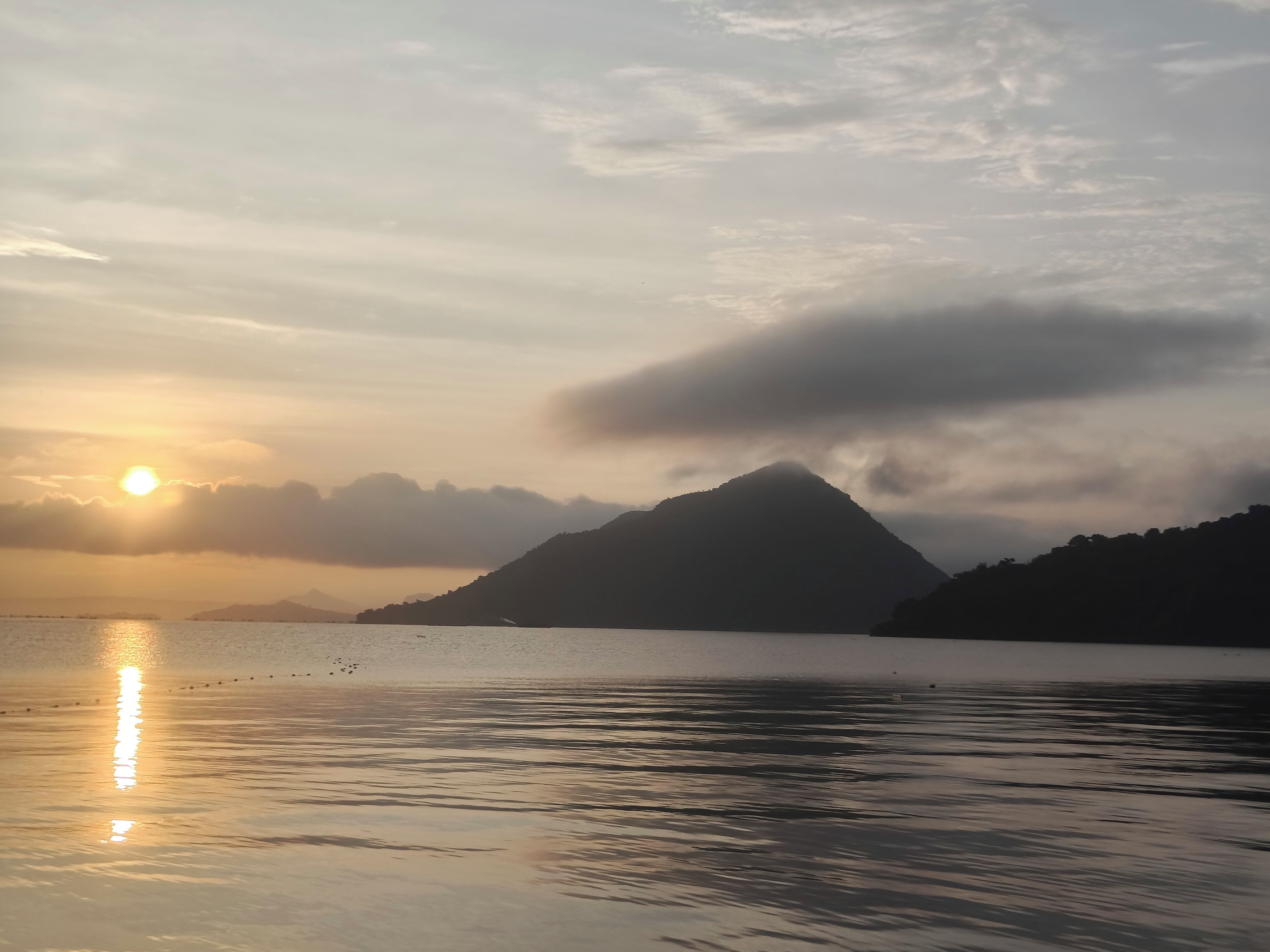 Une étendue d’eau calme où se reflète le soleil, avec une montagne en arrière-plan et un ciel nuageux.