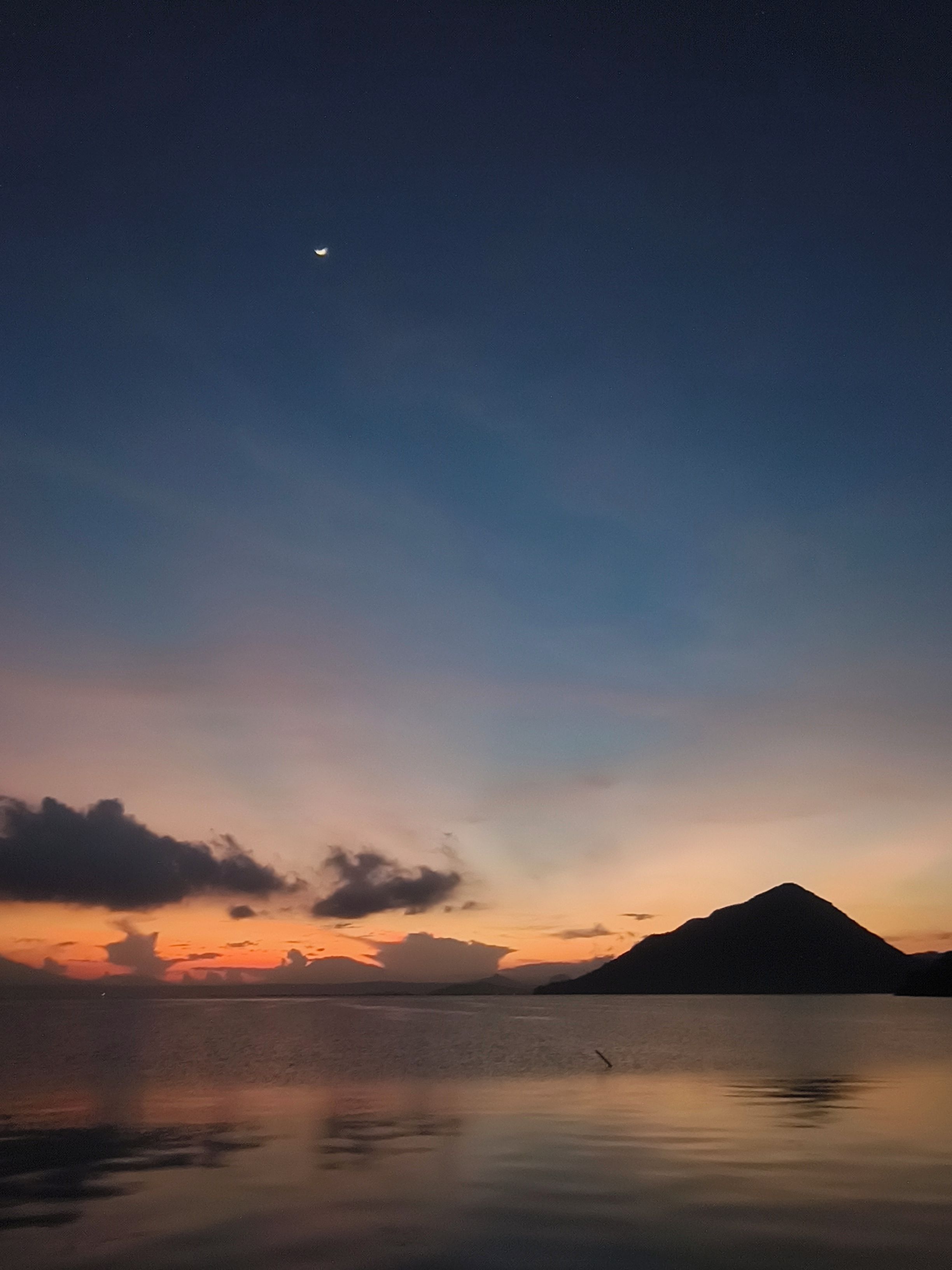 Acqua calma che riflette il cielo, la sagoma di una montagna e una luna crescente.